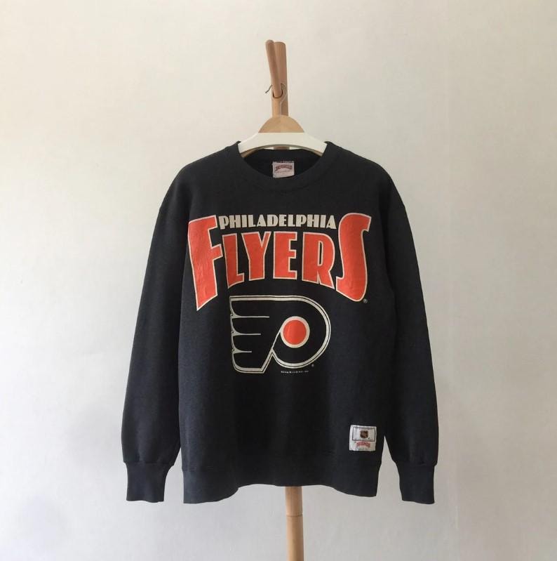 Może przedstawiać: Ciemnoszara bluza Philadelphia Flyers z pomarańczowym napisem i logo. Słowo "FLYERS" jest dużymi pomarańczowymi literami nad logo drużyny. Logo drużyny to stylizowane pomarańczowe koło w czarno-białym wzorze.