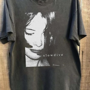 May include: Dark gray t-shirt featuring a black and white image of a woman's face with the band name "slowdive" printed in white. The shirt has short sleeves and a crew neck.