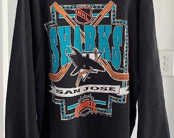 May include: Black sweatshirt featuring a teal and orange graphic of the San Jose Sharks hockey team logo. The logo includes the word "SHARKS" and a shark with a hockey stick. The NHL logo is also visible.
