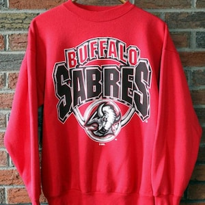May include: A red crew neck sweatshirt featuring the Buffalo Sabres logo. The logo includes the team name in black and white, with a buffalo head graphic. The sweatshirt is displayed against a brick wall.