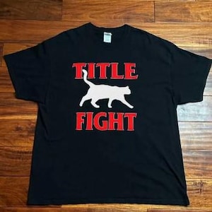 May include: Black t-shirt with the words "TITLE FIGHT" in red block letters above and below a white cat silhouette. The shirt is laid flat on a wooden surface.