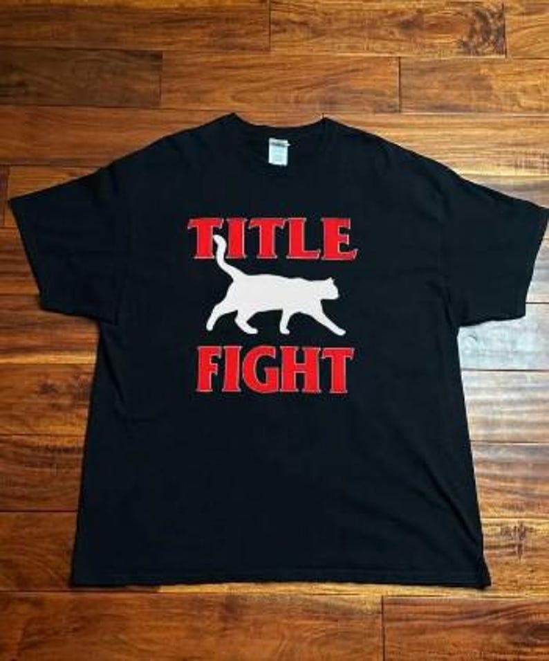 May include: Black t-shirt with the words "TITLE FIGHT" in red block letters above and below a white cat silhouette. The shirt is laid flat on a wooden surface.