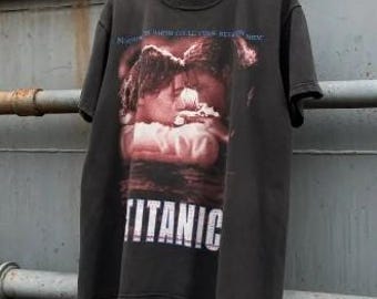 May include: Black t-shirt featuring a movie poster design from the film "Titanic." The graphic shows a romantic scene with the word "TITANIC" in white block letters. The shirt is hanging on a white hanger.