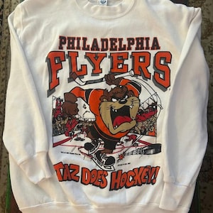 May include: White crew neck sweatshirt featuring the Philadelphia Flyers logo and Taz from Looney Tunes playing hockey. The graphic includes the text "TAZ DOES HOCKEY!" in orange and black. The sweatshirt has long sleeves and a classic fit.