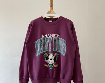May include: A maroon sweatshirt with the words "Anaheim Mighty Ducks" in white and teal lettering. The sweatshirt features the team's logo, a hockey mask and crossed hockey sticks, in teal, white, and gold.