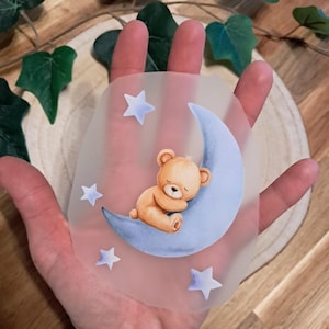 Könnte beinhalten: Ein durchscheinendes Transferblatt mit einem hellbraunen Teddybären, der auf einem weichen blauen Halbmond schläft, umgeben von vier kleinen, hellblauen Sternen. Das Motiv ist auf ein klares, flexibles Material mit matter Oberfläche gedruckt.