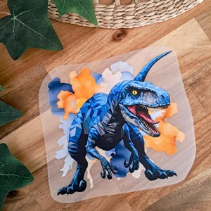 Könnte beinhalten: Ein blaues geometrisches Design eines Dinosauriers mit offenem Maul, vor einem Aquarellhintergrund in Orange- und Blautönen. Das Bild befindet sich auf einem transparenten, rechteckigen Aufkleber.