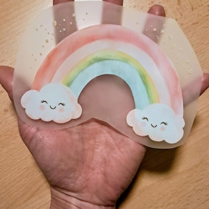 Könnte beinhalten: Ein Regenbogen-Aufkleber im Aquarellstil mit einer lächelnden Wolke an jedem Ende. Der Regenbogen zeigt Farbtöne von Rot, Orange, Gelb, Grün und Blau. Der Aufkleber wird in einer Hand gehalten und hat einen durchscheinenden Hintergrund mit Goldflocken.