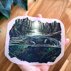 Könnte beinhalten: Ein rechteckiger Aufkleber mit einer detaillierten Illustration eines Alligators, der teilweise in einem dunklen Sumpf untergetaucht ist. Die Szene zeigt das Reptil, wie es durch trübes Wasser schwimmt, umgeben von dichten Zypressen und Laub in Dunkelgrün- und Brauntönen.