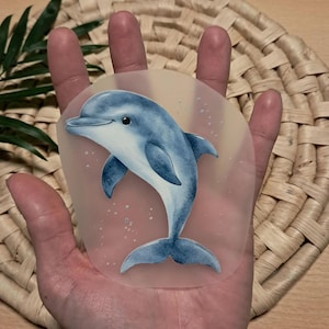 Könnte beinhalten: Ein blau-weißer Delfin-Aufkleber mit Aquarell-Effekt. Der Delfin wird beim Schwimmen dargestellt, mit einem gebogenen Körper und einem lächelnden Gesicht. Der Aufkleber befindet sich auf einem transparenten Hintergrund mit kleinen weißen Blasen.
