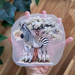 Könnte beinhalten: Ein dekorativer Aufkleber mit einem Zebra, das neben einem Baum steht. Das Zebra hat schwarz-weiße Streifen. Der Hintergrund ist ein zarter Rosaton mit einem Baum und Gras. Der Aufkleber wird in einer Hand gehalten.