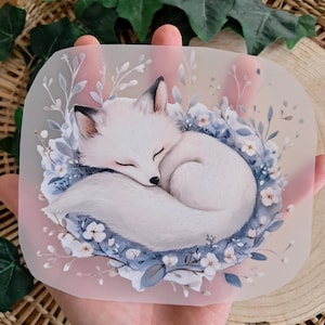 Könnte beinhalten: Ein rechteckiger, durchscheinender Aufkleber mit einem schlafenden weißen Fuchs, der sich in einem Bett aus blauen und weißen Blumen und Blättern zusammenrollt. Der Fuchs hat schwarzspitzige Ohren und ein geschlossenes Auge, mit einem weichen rosa Hintergrund.