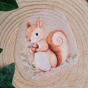 Könnte beinhalten: Aquarellillustration eines niedlichen Eichhörnchens, das eine Eichel hält. Das Eichhörnchen hat einen flauschigen Schwanz und ist von grünen Blättern umgeben. Das Kunstwerk befindet sich auf einer Holzscheibe.