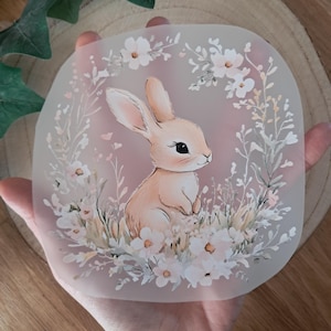 Könnte beinhalten: Quadratisches Bild mit einer pastellfarbenen Hasenillustration, umgeben von weißen Blumen und Blättern. Der Hase ist hellbraun mit langen Ohren und einem sanften Ausdruck. Das Bild wird in einer Hand gehalten, mit einem Holzhintergrund.