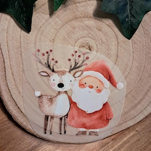 Könnte beinhalten: Aquarellillustration von Weihnachtsmann und Rentier. Der Weihnachtsmann trägt einen roten Anzug und Hut mit weißem Bart. Das Rentier hat ein Geweih mit roten Beeren. Das Bild befindet sich auf einem Holzgrund mit grünen Blättern.