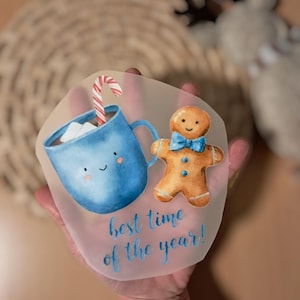 Könnte beinhalten: Aquarell-Illustration mit einer blauen Tasse mit Marshmallows und einem Zuckerstangen, sowie einem Lebkuchenmann mit blauer Fliege. Der Text "best time of the year!" ist in blauer Schrift geschrieben.