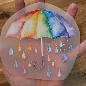 Könnte beinhalten: Ein durchscheinender Aufkleber mit einer Aquarellillustration eines regenbogenfarbenen Regenschirms mit violettem Griff. Das Design zeigt verschiedene bunte Regentropfen, die unter dem Schirm fallen, in Blau-, Rosa- und Gelbtönen.
