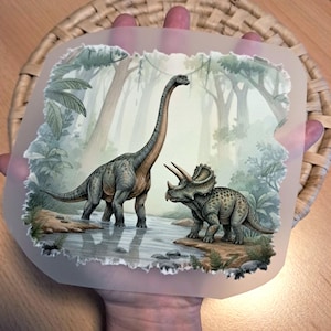 Könnte beinhalten: Illustration mit einem Brachiosaurus und einem Triceratops in einer prähistorischen Szene. Die Dinosaurier sind in Grau- und Grüntönen dargestellt und stehen in der Nähe von Wasser. Der Hintergrund zeigt Bäume und Blätter, was eine üppige, grüne Umgebung schafft.