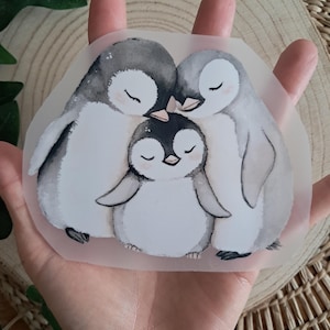 Könnte beinhalten: Ein Aufkleber mit einer Aquarellillustration von drei Pinguinen. Die Pinguine umarmen sich als Familie, wobei die Elternpinguine zu beiden Seiten eines Baby-Pinguins stehen. Die Pinguine sind hauptsächlich weiß, grau und schwarz, mit geschlossenen Augen.