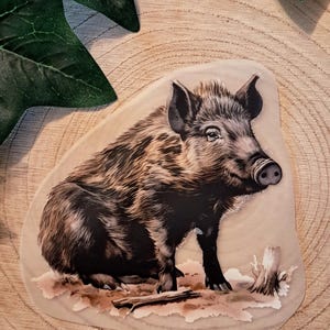 Könnte beinhalten: Ein Aufkleber mit einer detaillierten Illustration eines Wildschweins. Das Wildschwein ist in Braun- und Schwarztönen dargestellt, mit einem strukturierten Fell und einer markanten Schnauze. Der Aufkleber hat einen hellbeigen Hintergrund und befindet sich auf einer Holzoberfläche.