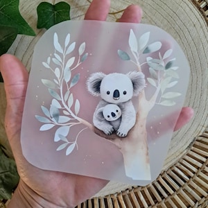Könnte beinhalten: Ein klarer, quadratischer Untersetzer mit einer Aquarellillustration eines Koala-Muttertiers und eines Baby-Koalas in einem Baum. Das Design umfasst weiße und graue Blätter und Zweige mit einem weichen, rosafarbenen Hintergrund.