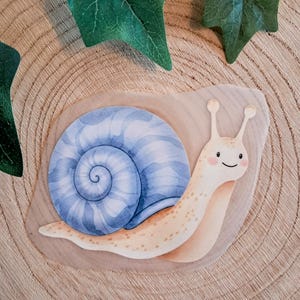 Könnte beinhalten: Ein hölzerner Schnecken-Ausschnitt mit einem hellblau-weiß gewirbelten Gehäuse. Der Schneckenkörper ist hellbeige mit einem lächelnden Gesicht. Der Ausschnitt befindet sich auf einer Holzoberfläche mit grünen Efeublättern.