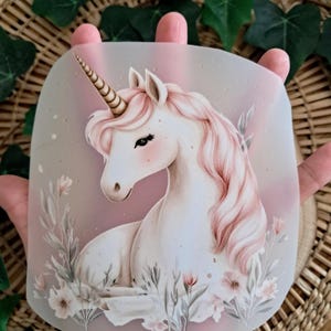 Könnte beinhalten: Ein pastellfarbenes Einhorn-Design. Das Einhorn hat einen weißen Körper, eine rosa Mähne und ein goldenes Horn. Zarte Blumen- und Blattelemente umgeben das Einhorn. Das Bild ist vor einem weichen, farbverlaufenden Hintergrund.
