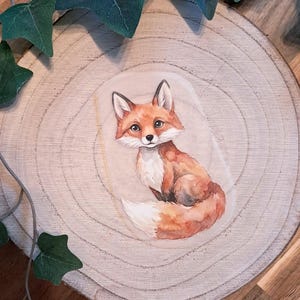 Könnte beinhalten: Aquarellillustration eines Fuchses mit orangefarbenem, braunem und weißem Fell. Der Fuchs sitzt auf einer hellen ovalen Form. Der Hintergrund ist eine Holzscheibe mit grünen Blättern.