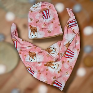 Könnte beinhalten: Ein rosa Baby-Mütze und passendes Lätzchen-Set. Das Set zeigt ein sich wiederholendes Muster aus Popcorn, Brezeln und Milchshakes. Mütze und Lätzchen sind aus weichem Stoff, ideal für ein Baby.