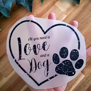 Könnte beinhalten: Ein herzförmiges Design mit dem Text "All you need is Love and a Dog" und einem schwarzen Pfotenabdruck. Das Design ist in schwarzer Schrift auf einem durchscheinenden Hintergrund.