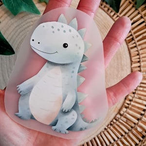 Könnte beinhalten: Ein hellblauer Dinosaurier-Aufkleber mit freundlichem Gesichtsausdruck. Der Aufkleber hat einen weißen Bauch und einen rosa Hintergrund. Der Dinosaurier hat eine Reihe grüner Stacheln auf dem Rücken. Der Aufkleber wird in einer Hand gehalten.