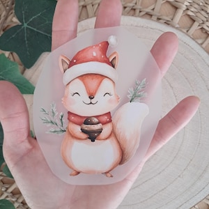 Könnte beinhalten: Eine Illustration eines Eichhörnchens mit Weihnachtsmütze und rotem Schal, das eine Eichel hält. Das Eichhörnchen hat einen buschigen Schwanz und ist von Tannenzweigen umgeben. Das Kunstwerk befindet sich auf einem transparenten, runden Aufkleber.