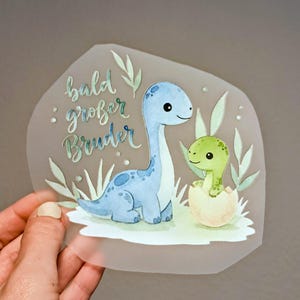 Könnte beinhalten: Aquarell-Illustration von zwei Dinosauriern auf transparentem Hintergrund. Ein blauer Dinosaurier steht neben einem grünen Dinosaurier, der aus einem Ei schlüpft. Der Text "bald großer Bruder" ist in Schrift geschrieben.