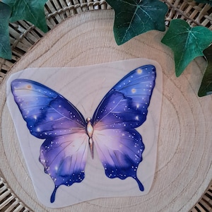 Könnte beinhalten: Eine Holzdekoration in Form eines Schmetterlings mit einem Aquarelldesign. Der Schmetterling hat blaue und violette Flügel mit weißen sternartigen Akzenten. Die Dekoration befindet sich auf einer Holzscheibe mit gewebtem Hintergrund und grünen Blättern.