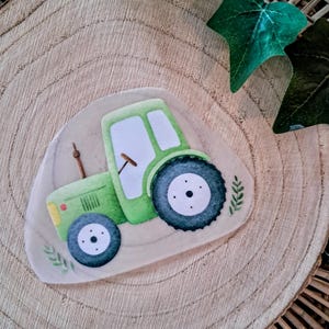 Könnte beinhalten: Eine grüne Traktorillustration mit schwarzen Reifen und einer weißen Kabine ist auf einem hellbraunen, oval geformten Holzstück zentriert. Der Traktor hat ein Lenkrad und einen Schornstein. Der Hintergrund ist eine Holzoberfläche mit grünen Blättern.
