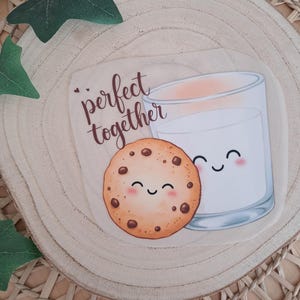 Könnte beinhalten: Eine verspielte Illustration eines Schokoladenkekses und eines Glases Milch, beide mit lächelnden Gesichtern, mit der Aufschrift "perfect together". Das Bild befindet sich auf einer Holzscheibe und erzeugt eine gemütliche Atmosphäre.