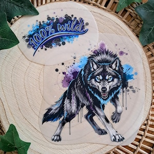 Könnte beinhalten: Zwei Holzuntersetzer mit einer Wolfsillustration und dem Text "100% wild". Der Wolf ist in Schwarz, Blau und Lila dargestellt, in einer aggressiven Haltung. Die Untersetzer liegen auf einer Holzoberfläche mit grünen Blättern.