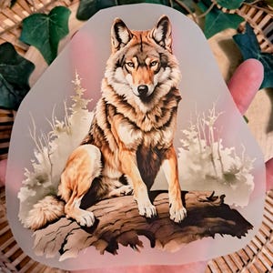 Könnte beinhalten: Ein durchscheinendes, unregelmäßig geformtes Dekorationsteil mit einer detaillierten Illustration eines Wolfes. Der Wolf hat braunes und hellbraunes Fell und sitzt auf einem Baumstamm in einer Waldlandschaft. Der Hintergrund umfasst stilisierte Bäume und einen weichen, rosafarbenen Farbton.
