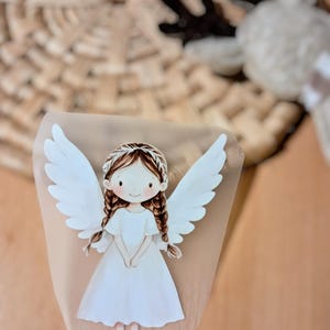 Könnte beinhalten: Ein Papierengel mit weißen Flügeln und einem weißen Kleid. Der Engel hat braunes geflochtenes Haar und ein lächelndes Gesicht. Der Hintergrund ist hellbraun.