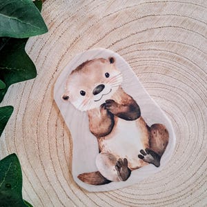 Könnte beinhalten: Ein Aufkleber mit einer Aquarellillustration eines braunen und cremefarbenen Seeotters mit lächelndem Ausdruck, dunklen Augen und rosa Wangen, der in sitzender Position dargestellt ist und seine Pfoten nahe an der Brust hält.