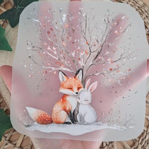 Könnte beinhalten: Ein klares, quadratisches Dekorationsteil mit einer Aquarellillustration eines Fuchses und eines Kaninchens, die sich unter einem Baum mit rosa Blüten umarmen. Der Fuchs ist orange und weiß, und das Kaninchen ist weiß. Der Hintergrund ist ein zartes Rosa.