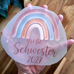 Könnte beinhalten: Ein durchscheinender, halbkreisförmiger Aufkleber mit einem Regenbogen im Aquarellstil in Blau-, Pfirsich- und Rosatönen. Unter dem Regenbogen steht der Text "große Schwester 2027" in einer dunklen Schreibschrift, verziert mit kleinen Sternen.