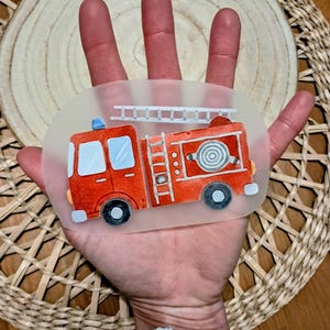 Könnte beinhalten: Ovaler Aufkleber mit einer rot-weißen Feuerwehrwagen-Illustration. Das Design umfasst eine Leiter, einen Schlauch und ein blaues Licht. Der Feuerwehrwagen hat schwarze Räder und einen transparenten Hintergrund.