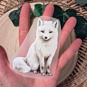 Könnte beinhalten: Ein Aufkleber mit einem weißen Polarfuchs, mit einer detaillierten Illustration. Der Fuchs sitzt, mit seinem flauschigen Schwanz eingerollt. Der Aufkleber wird in einer Hand gehalten, mit einem natürlichen Hintergrund aus Holz und Grün.