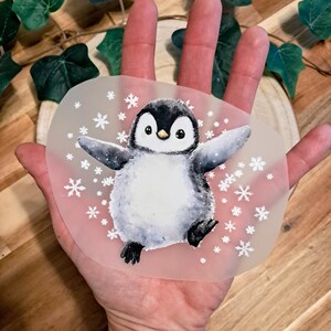 Könnte beinhalten: Aquarell-Illustration eines Pinguins mit ausgebreiteten Flügeln, umgeben von weißen Schneeflocken. Der Pinguin ist hauptsächlich schwarz und weiß mit einem gelben Schnabel, vor einem durchscheinenden Hintergrund. Das Bild wird in einer Hand gehalten.
