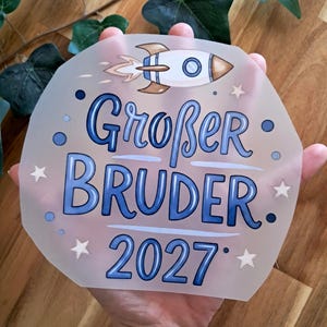 Könnte beinhalten: Ein durchscheinendes, sechseckiges Schild mit den Worten "Großer Bruder 2027" in blauer Schrift, zusammen mit einer Raketenillustration. Das Design umfasst kleine Sterne und Punkte. Das Schild wird von der Hand einer Person gehalten.