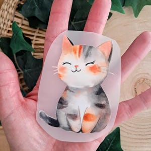 Könnte beinhalten: Ein Aufkleber im Aquarellstil mit einer lächelnden Katze mit orangefarbenem, grauem und weißem Fell. Die Katze hat rosige Wangen und einen schwarzen Schwanz. Der Aufkleber wird in einer Hand gehalten, mit einem geflochtenen Korb und grünen Blättern im Hintergrund.