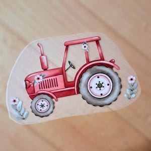 Könnte beinhalten: Ein flacher, hellfarbener Holzausschnitt mit einer Illustration eines roten Traktors. Der Traktor hat große dunkelgraue Reifen, ein Lenkrad und einen Schornstein. Kleine rosa und weiße Blumenakzente befinden sich auf dem Traktor.