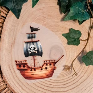 Könnte beinhalten: Ein Piratenschiff-Aufkleber mit braunem Rumpf, schwarz-weiß gestreiften Segeln und einem Totenkopf-Emblem. Das Schiff hat eine schwarze Flagge und ist vor einem hellen Hintergrund platziert. Der Aufkleber ist von grünen Blättern umgeben.