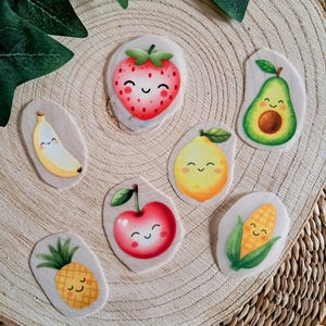 Könnte beinhalten: Eine Sammlung von Holzmagneten in Form von Früchten, darunter Erdbeere, Banane, Avocado, Zitrone, Kirsche, Ananas und Mais. Jeder Magnet zeigt ein lächelndes Gesicht und ist in hellen, fröhlichen Farben bemalt. Auf einer Holzoberfläche angeordnet.